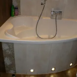 Jasna, narożna wanna z hydromasażem, obudowana płytkami w odcieniach beżu i brązu, z oświetleniem punktowym u dołu, świeczka zapalona na brzegu wanny.