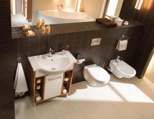 Nowoczesna łazienka z brązowymi płytkami, białą umywalką z szafką, toaletą i bidetem, uzupełniona o akcesoria łazienkowe i oświetlenie.