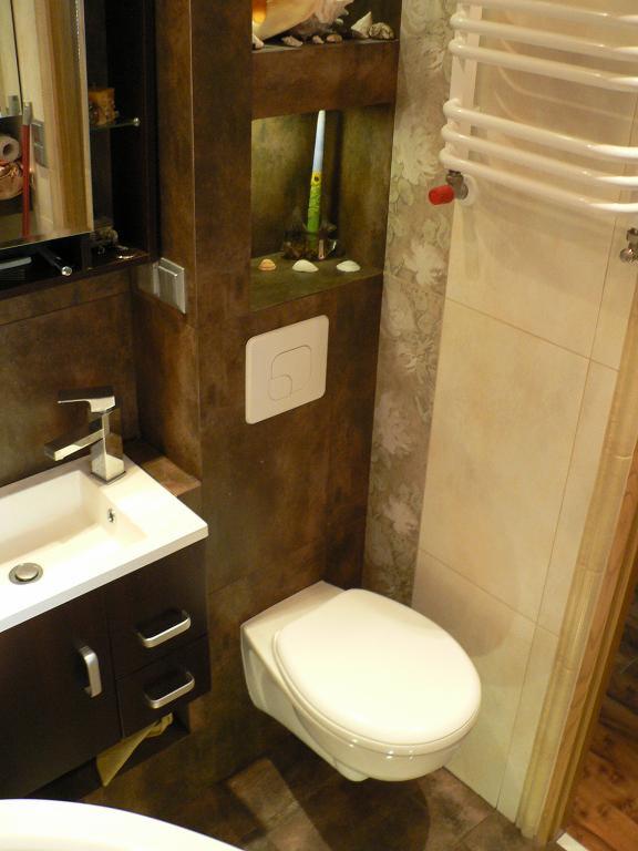 Nowoczesna łazienka z brązowymi płytkami, wiszącą toaletą, umywalką z szafką i dekoracyjną półką z oświetleniem oraz grzejnikiem drabinkowym.