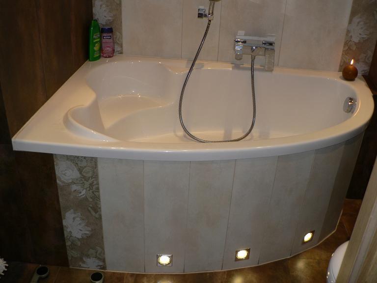 Jasna, narożna wanna z hydromasażem, obudowana płytkami w odcieniach beżu i brązu, z oświetleniem punktowym u dołu, świeczka zapalona na brzegu wanny.