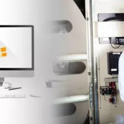 Uśmiechnięty mężczyzna z identyfikatorem, pracujący na laptopie przed otwartą szafą sterowniczą z widocznymi bezpiecznikami i okablowaniem; po lewej stronie biurko z komputerem iMac wyświetlającym...