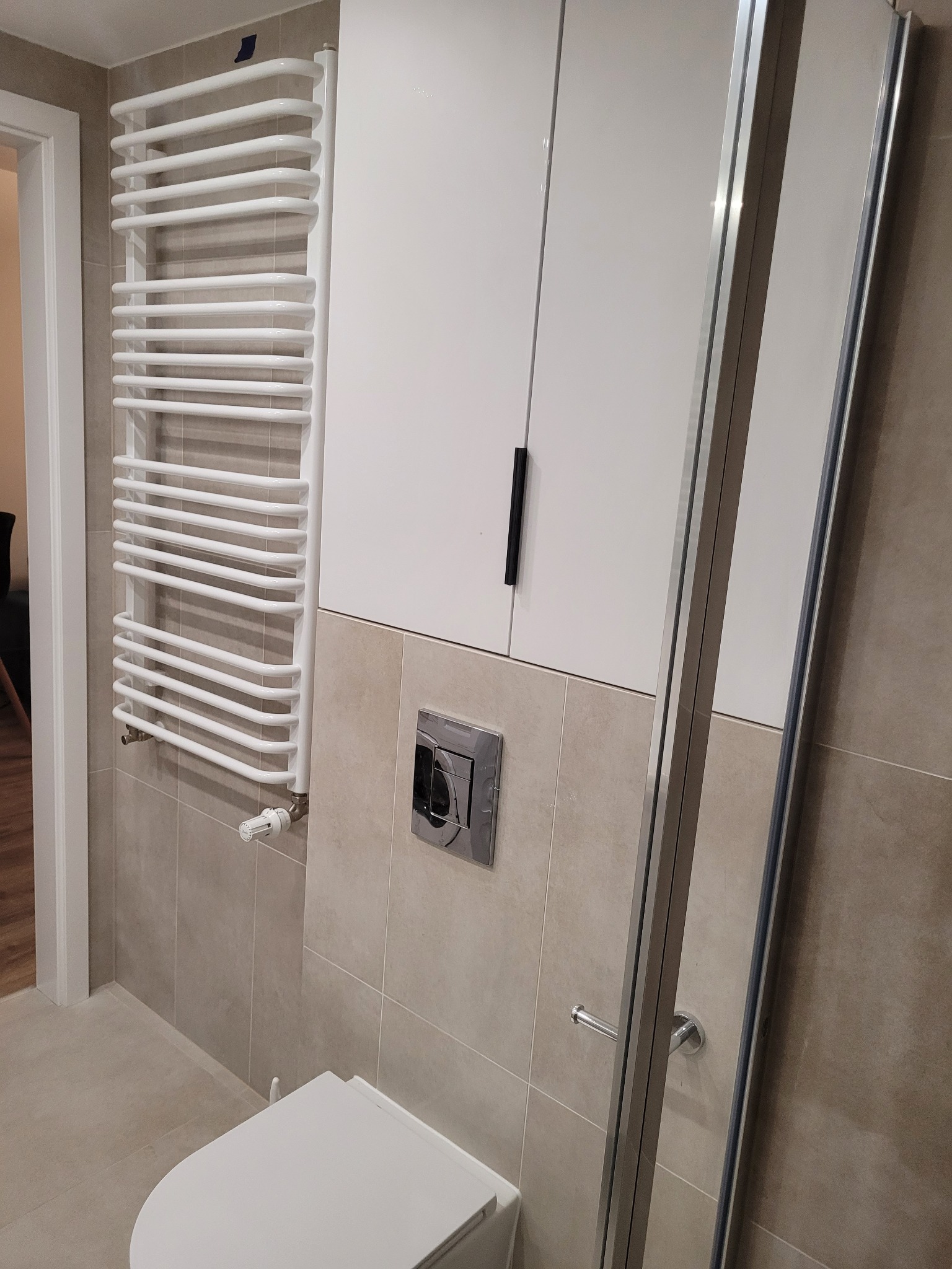 Wykończona łazienka z beżowymi płytkami, białym grzejnikiem drabinkowym, zabudową meblową z szafkami i kabiną prysznicową z chromowanymi profilami.