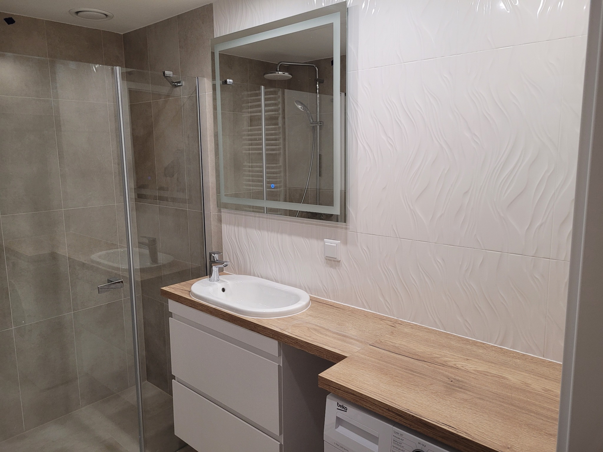 Nowoczesna łazienka z kabiną prysznicową, umywalką nablatową na drewnianym blacie, białe płytki z falistym wzorem oraz podświetlane lustro z widocznym odbiciem słuchawki prysznicowej.