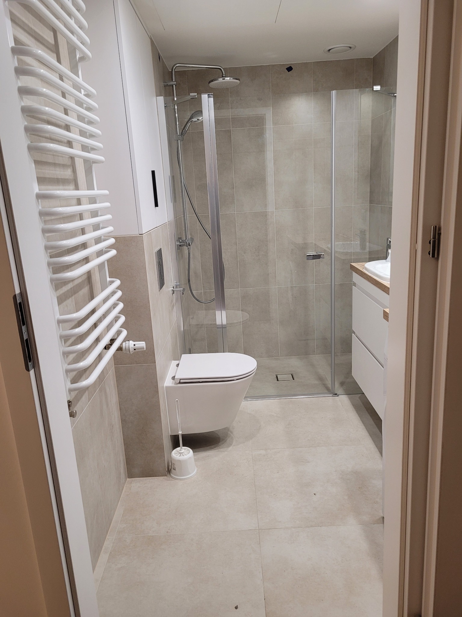 Wykończona łazienka z kabiną prysznicową, toaletą i grzejnikiem drabinkowym, płytki w odcieniach beżu, widok zza futryny drzwi.