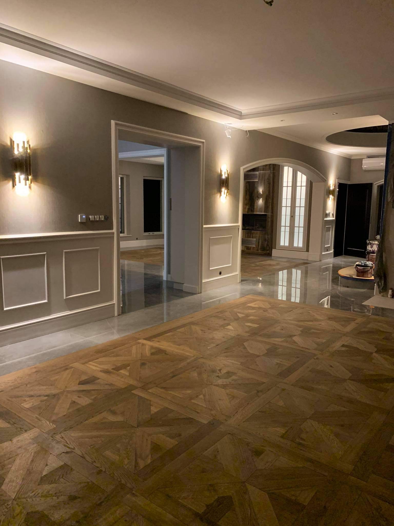 Wnętrze domu z drewnianą podłogą parkietową w geometryczny wzór, szare ściany z białymi panelami dekoracyjnymi, oświetlenie ścienne i widoczne przejścia do innych pomieszczeń.