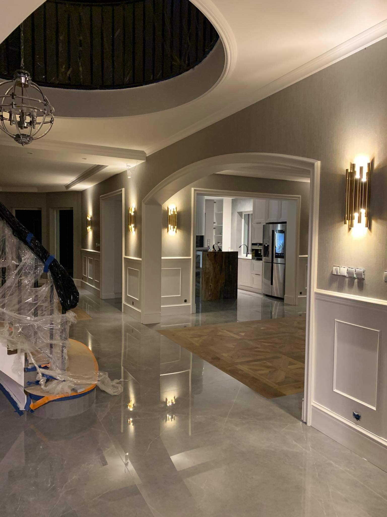 Hol w luksusowym domu z szarą marmurową podłogą, widoczne elementy dekoracyjne ścian, oświetlenie i częściowo zakryte schody w folii ochronnej.