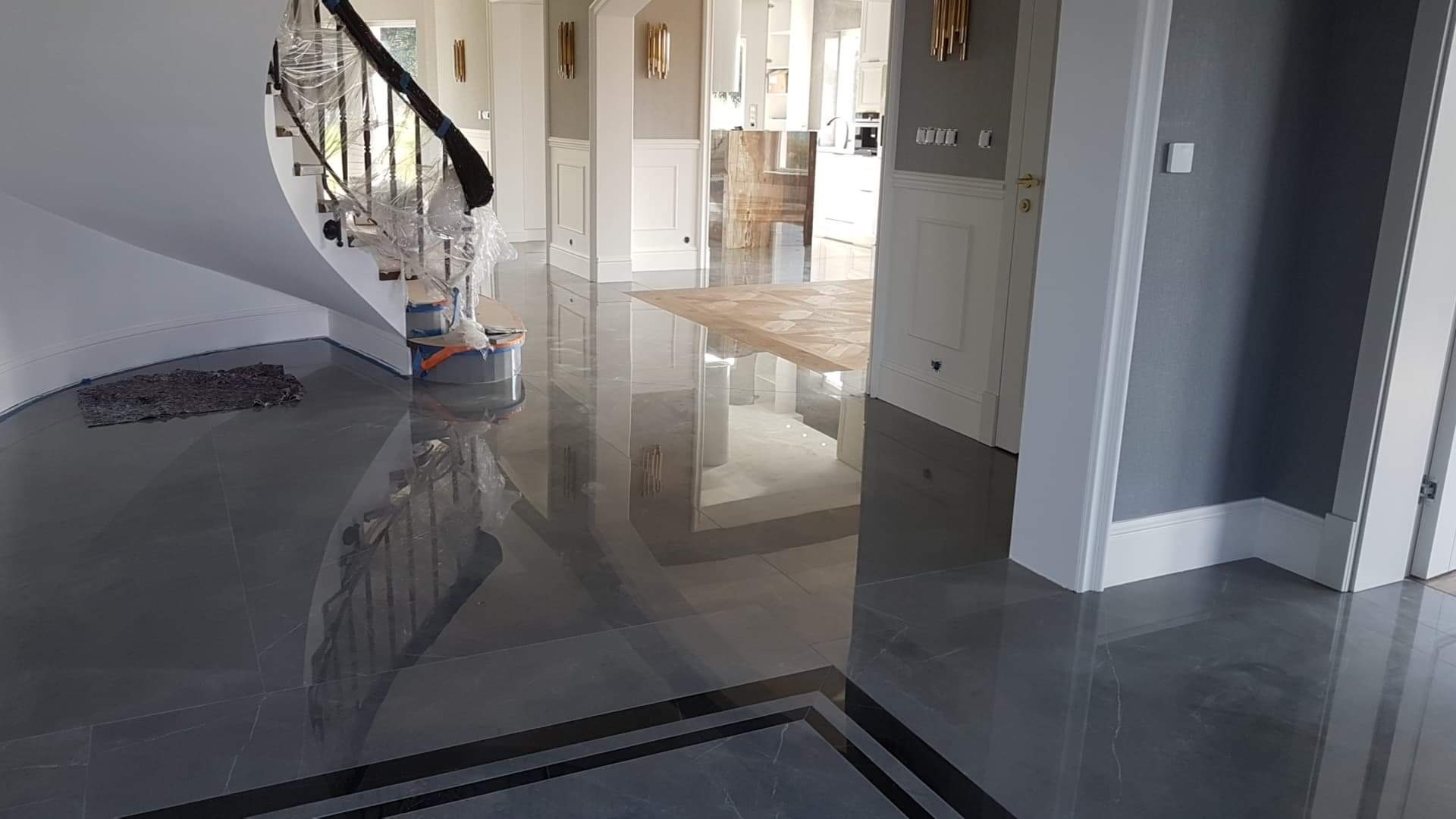 Polerowana, ciemnoszara podłoga z płytek z czarną bordiurą, odbijająca światło, z widocznymi schodami zabezpieczonymi folią i wiaderkiem z narzędziami w tle.