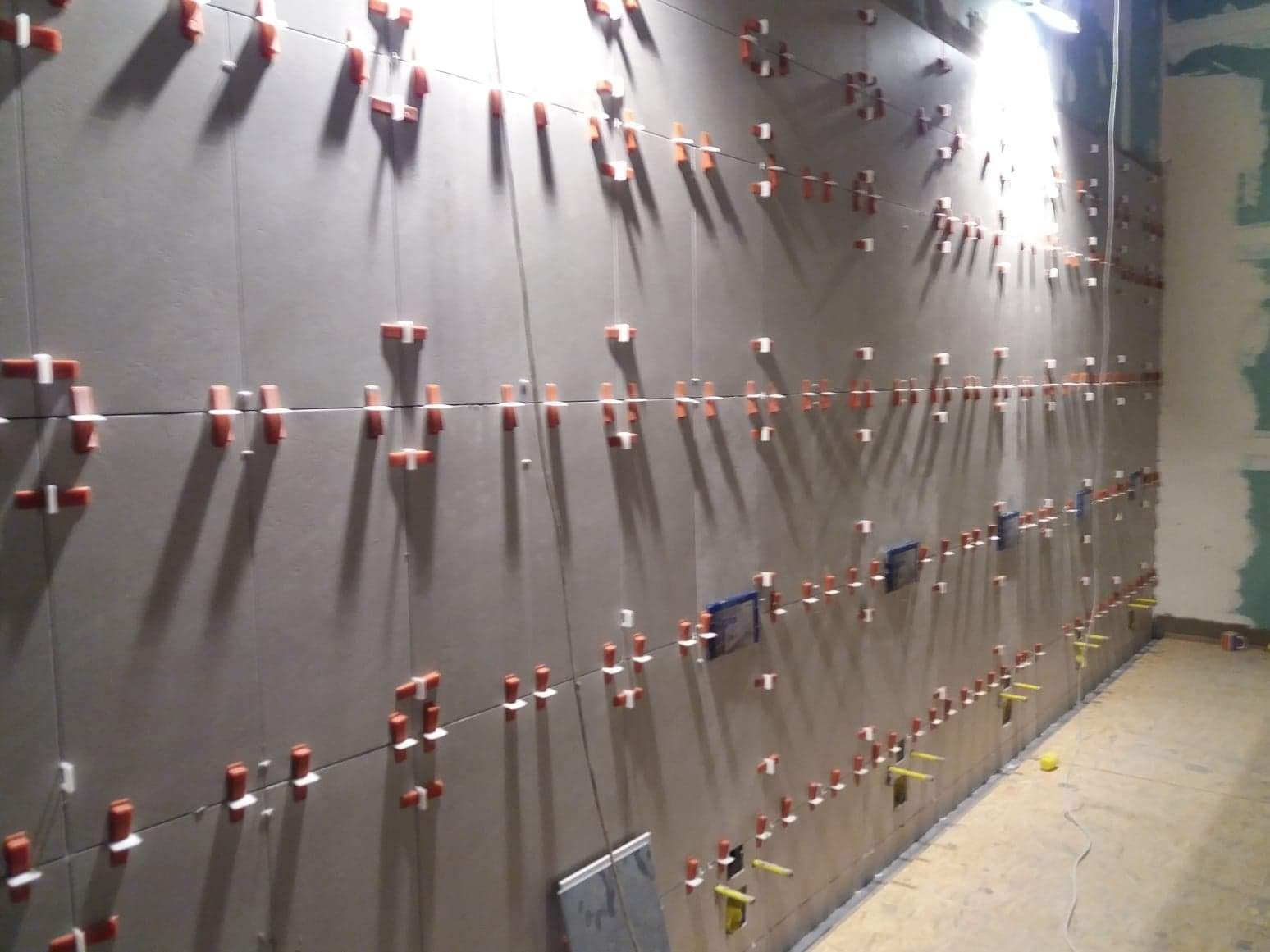 Ściana w trakcie układania płytek ceramicznych z użyciem systemów poziomowania, widoczne krzyżyki dystansowe i kliny, niedokończone prace wykończeniowe na sąsiedniej ścianie.