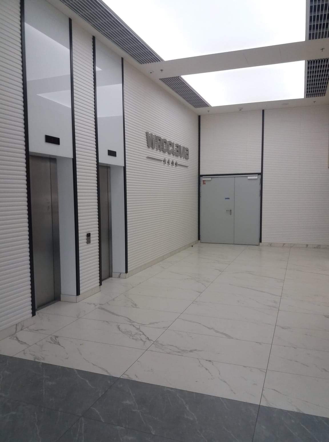 Wnętrze nowoczesnego biurowca z marmurową posadzką, dwoma windami w czarnych obramowaniach i napisem 'WROCLEVE' na ścianie z poziomymi żłobieniami.