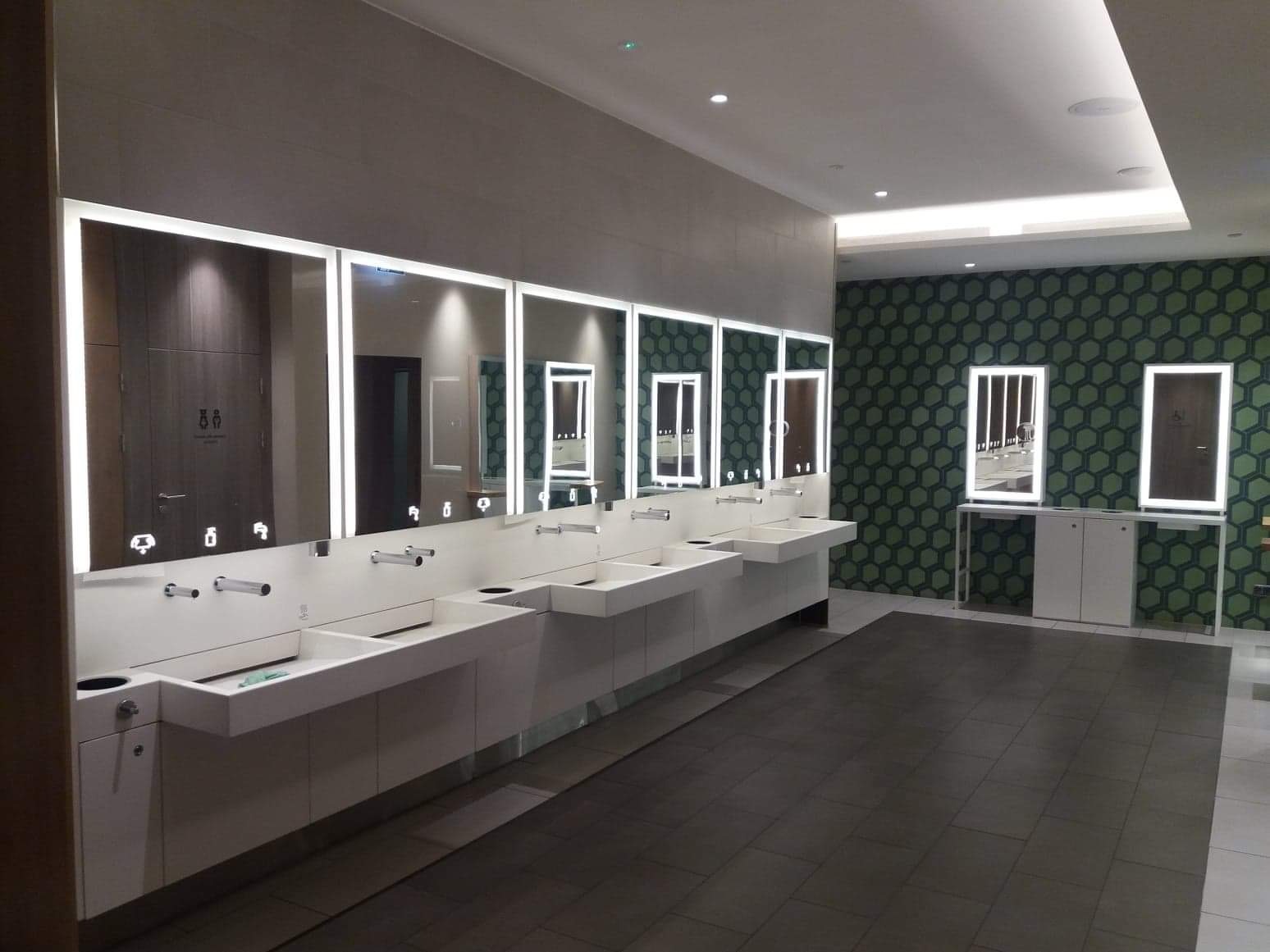 Nowoczesna toaleta publiczna z długim rzędem białych umywalek z podświetlanymi lustrami, ciemną podłogą i ścianą w geometryczny wzór.