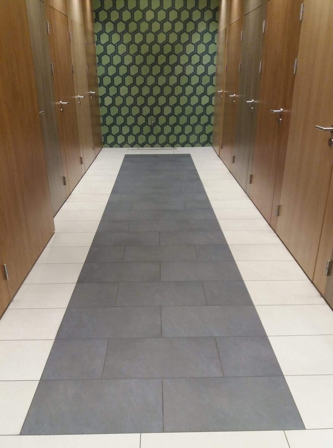 Wykończony korytarz z szarymi płytkami ułożonymi w pas na środku i białymi po bokach, otoczony drewnianymi szafkami i zieloną tapetą w geometryczne wzory.