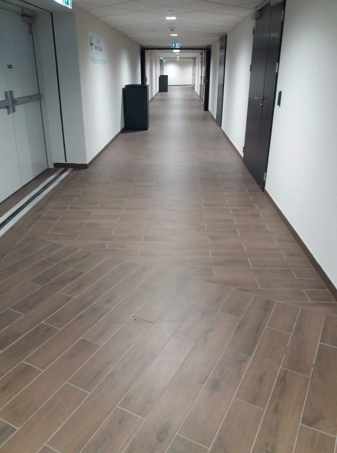 Długi korytarz wyłożony brązowymi płytkami imitującymi drewno, ułożonymi we wzór jodełki na środku, z widoczną małą skazą na jednej z płytek, prowadzący w kierunku jasnego światła na końcu.