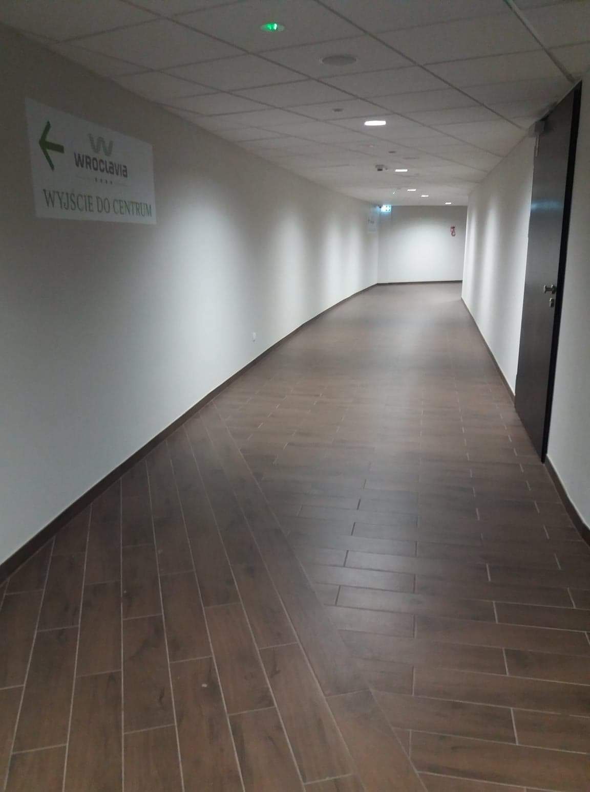 Długi korytarz z brązową podłogą ułożoną w jodełkę, białe ściany z oznaczeniem kierunku do centrum, oświetlenie sufitowe.