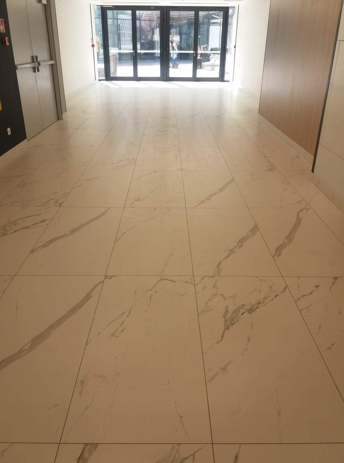 Beżowa podłoga z płytek imitujących marmur w nowoczesnym holu, widok w kierunku przeszklonych drzwi wejściowych z widocznymi sylwetkami osób.