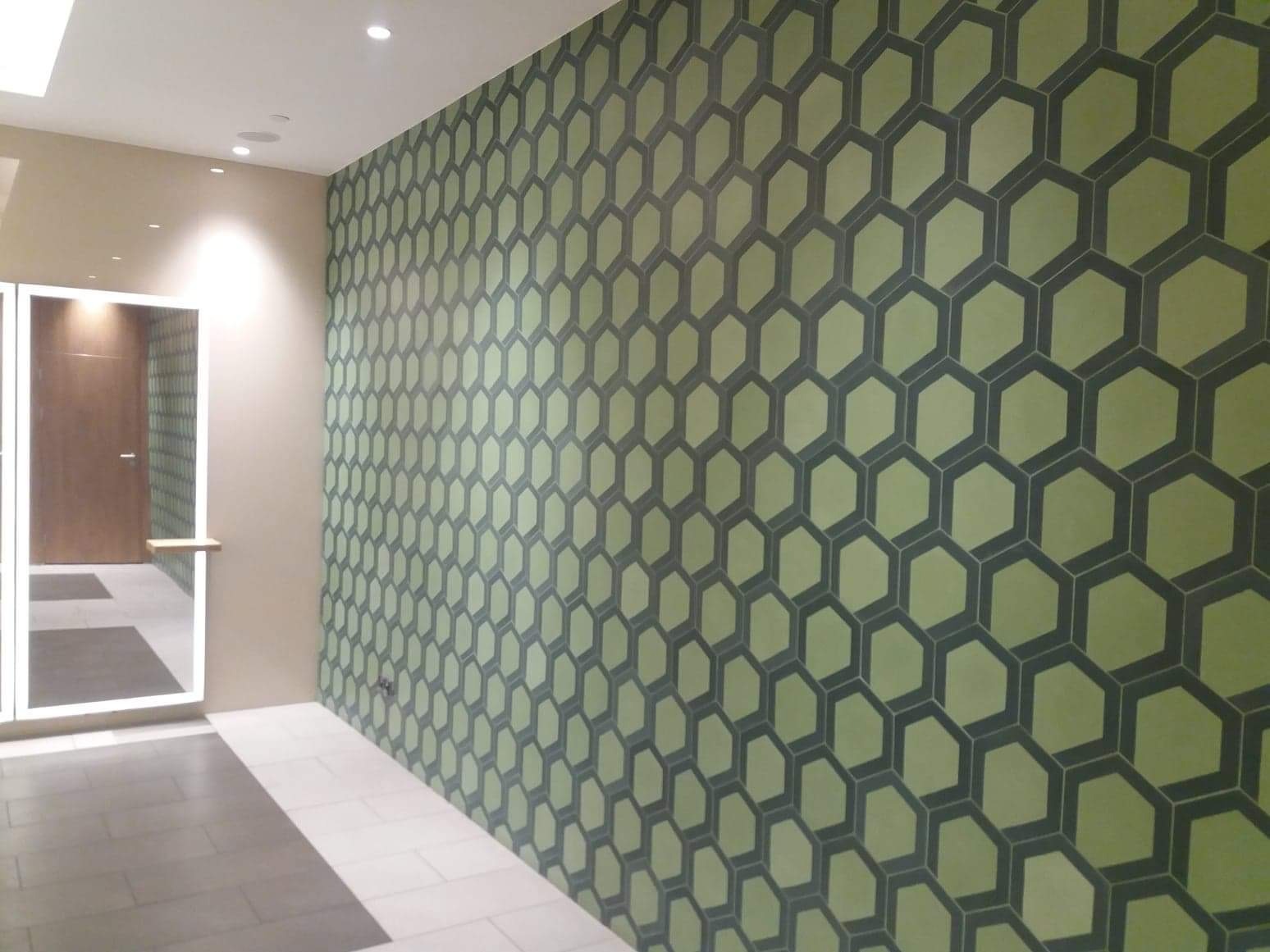 Ściana wyłożona heksagonalnymi płytkami w odcieniach zieleni i szarości, widoczna w korytarzu z minimalistycznym oświetleniem i wejściem do pomieszczenia z drewnianymi drzwiami.