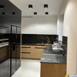 Nowoczesna kuchnia z drewnianymi frontami szafek, czarnym blatem z marmurkowym wzorem i białymi szafkami wiszącymi, oświetlona punktowymi lampami sufitowymi.