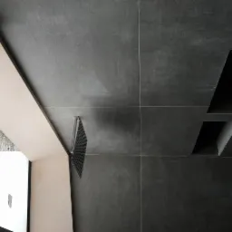 Ciemnoszare płytki ścienne z wnękami, widoczny fragment okna z białym obramowaniem i metalowa kratka wentylacyjna przy ścianie.