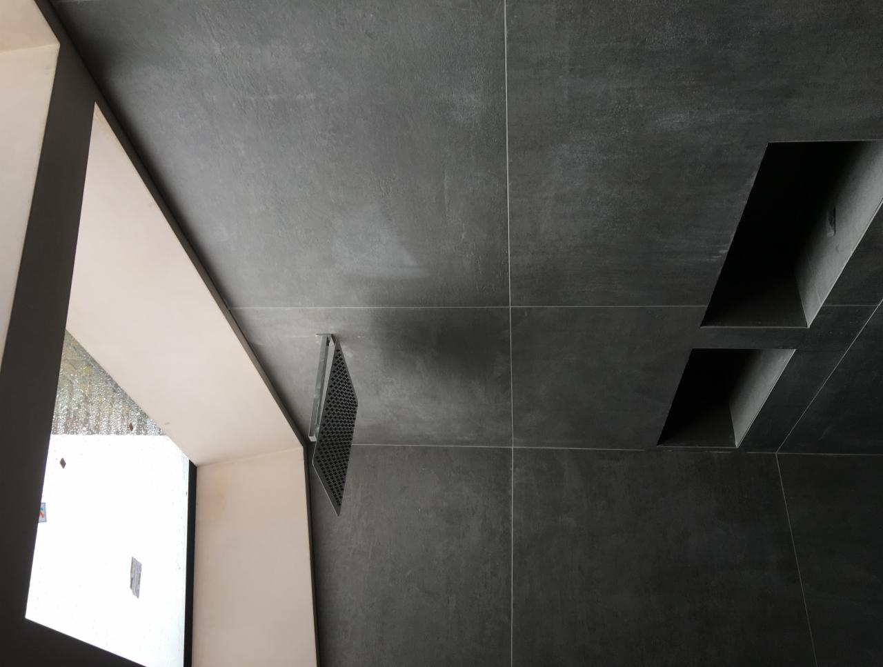 Ciemnoszare płytki ścienne z wnękami, widoczny fragment okna z białym obramowaniem i metalowa kratka wentylacyjna przy ścianie.