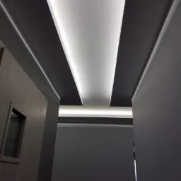 Nowoczesny sufit podwieszany z oświetleniem LED w kształcie pasów, szara kolorystyka ścian i sufitu, widoczny fragment drewnianych drzwi z dekoracyjnym wycięciem.