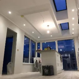 Wnętrze w trakcie odświeżania: świeżo pomalowane ściany w odcieniach szarości, widoczne okna dachowe, wyspa kuchenna z wyposażeniem i workiem na podłodze, oraz rozwieszone pranie w tle.