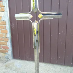 Krzyż wykonany ze stali nierdzewnej z polerowanymi elementami, ustawiony na betonowej powierzchni w Przysusze, na tle brązowej blaszanej ściany i fragmentu ceglanej ściany.