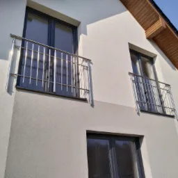 Elewacja domu z dwoma balkonami zabezpieczonymi nowoczesnymi, błyszczącymi balustradami ze stali nierdzewnej z pionowymi prętami, widoczne okna z szarymi ramami i drewniane wykończenie dachu.