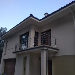 Dwukondygnacyjny dom z balkonem zabezpieczonym poziomą, metalową konstrukcją, widok z dołu na elewację w odcieniach beżu i brązu, z fragmentem ogrodu i drzew po lewej stronie.