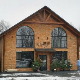 Usługi Projektowe Barbara Skupińska - Kurek - Dwukondygnacyjny budynek biurowy z cegły z dużymi łukowymi oknami i drewnianą konstrukcją dachu, oświetlony zimowym światłem, z napisem 'Biuro Ekspozycja' nad wejściem i choinkami w donicach przed...