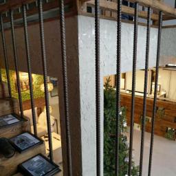 Usługi Projektowe Barbara Skupińska - Kurek - Schody z drewnianymi stopniami i metalową balustradą z prętów zbrojeniowych, na podeście ramki ze zdjęciami, w tle choinka i element wystroju wnętrza.