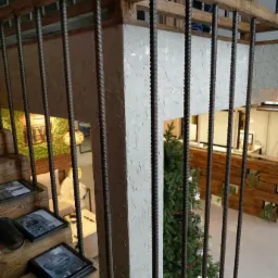 Schody z drewnianymi stopniami i metalową balustradą z prętów zbrojeniowych, na podeście ramki ze zdjęciami, w tle choinka i element wystroju wnętrza.