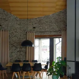 Wnętrze jadalni z ceglaną ścianą, drewnianym sufitem i dużymi oknami, oświetlone wiszącymi lampami w kształcie kopuł. Na pierwszym planie drewniany stół z czarnymi krzesłami i zielona roślina...