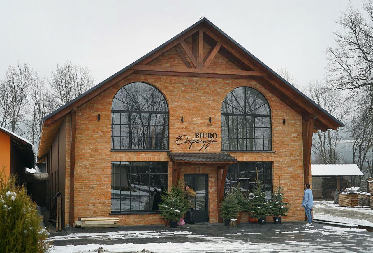 Dwukondygnacyjny budynek biurowy z cegły z dużymi łukowymi oknami i drewnianą konstrukcją dachu, oświetlony zimowym światłem, z napisem 'Biuro Ekspozycja' nad wejściem i choinkami w donicach przed...