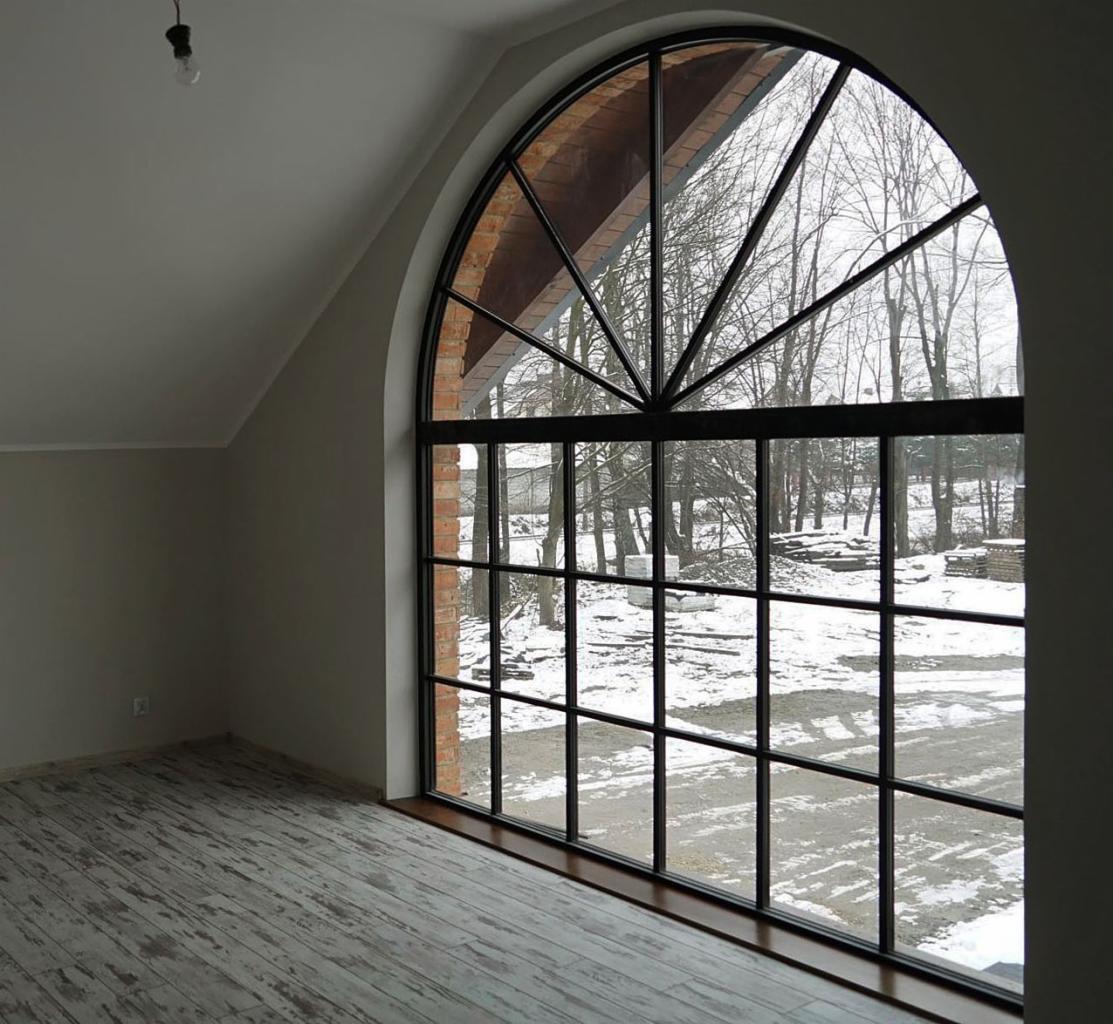 Duże, łukowe okno z czarnymi szprosami w surowym wnętrzu, widok na zimowy krajobraz z zewnątrz, ceglana wnęka okienna, drewniana podłoga z efektem postarzenia.
