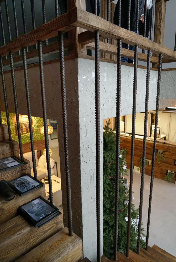 Schody z drewnianymi stopniami i metalową balustradą z prętów zbrojeniowych, na podeście ramki ze zdjęciami, w tle choinka i element wystroju wnętrza.