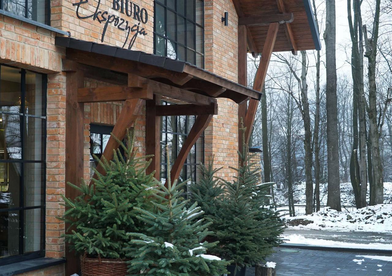 Wejście do biura z cegły z drewnianym zadaszeniem, ozdobione młodymi choinkami w donicach, zimowy krajobraz w tle.