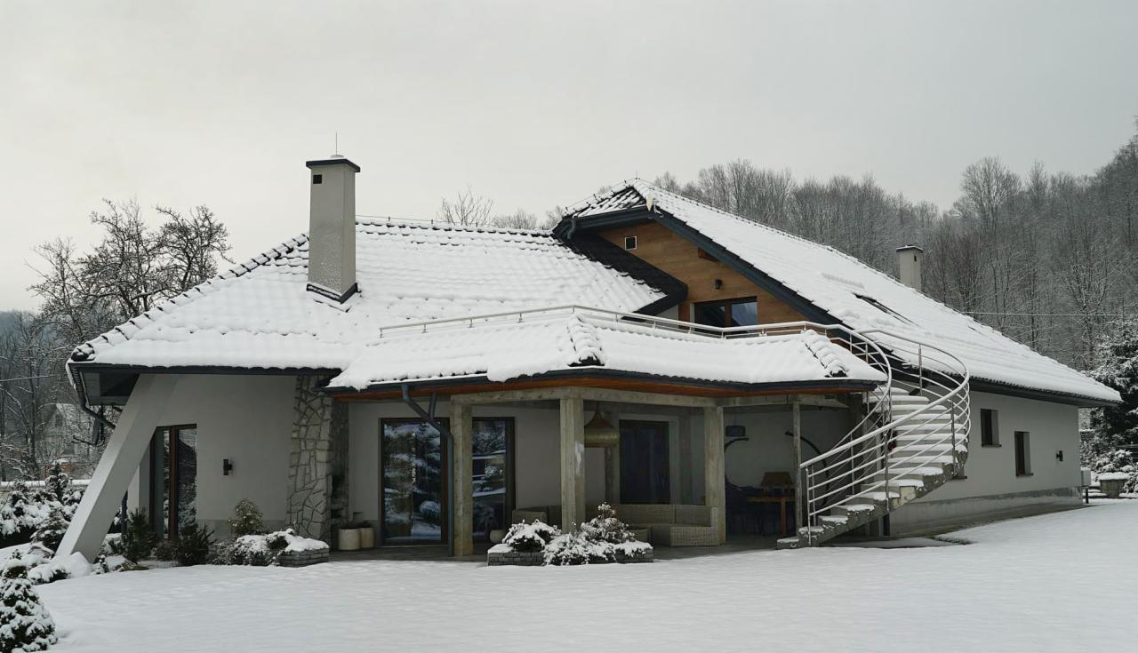 Nowoczesny dom jednorodzinny z białymi ścianami, pokryty śniegiem, z zewnętrznymi schodami spiralnymi i kamiennym wykończeniem elewacji.