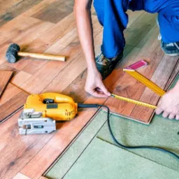 Pomiar panela podłogowego przed docięciem, widoczna żółta wyrzynarka, gumowy młotek i miarka. Pracownik w niebieskim kombinezonie podczas pracy.