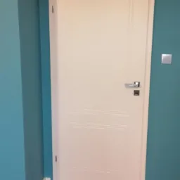 Białe drzwi z panelem w dolnej części i metalową klamką na tle turkusowej ściany, zamontowane w mieszkaniu w Krośnie.