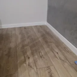 Ułożone panele podłogowe imitujące drewno, z widocznymi listwami przypodłogowymi przy białej i szarej ścianie.