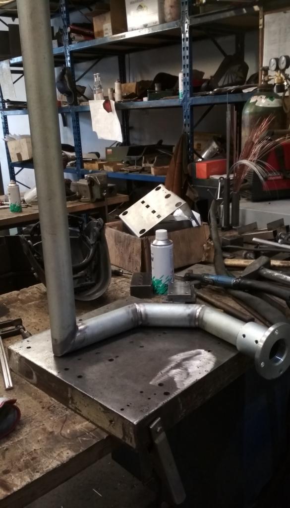 Spawany element rurowy z kołnierzem na metalowym stole warsztatowym, w tle regał z narzędziami i butla spawalnicza.