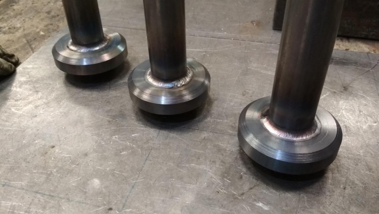 Trzy metalowe elementy z widocznymi spawami, leżące na metalowej powierzchni, z delikatnym odblaskiem światła na spawach.