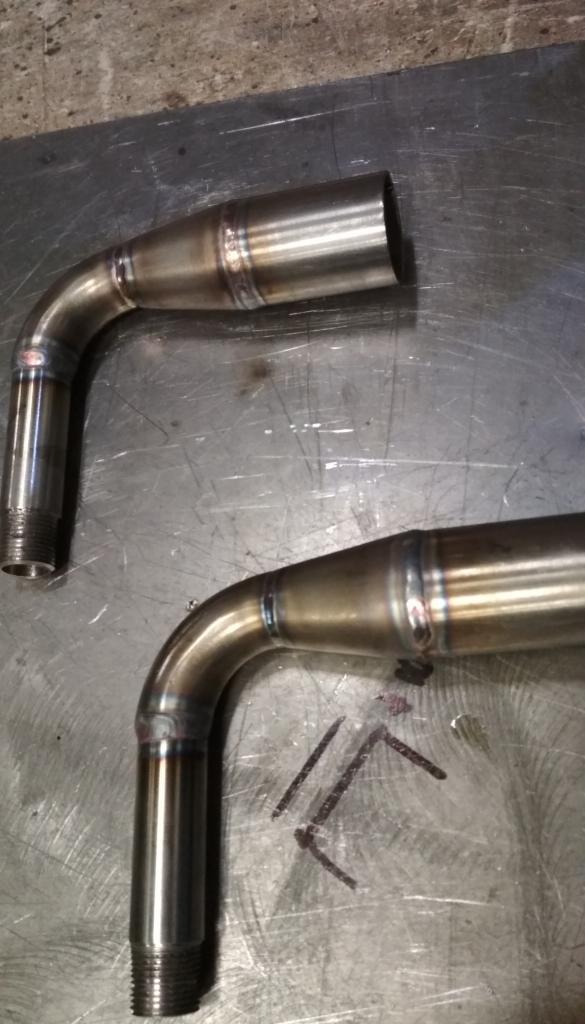 Dwa spawane, zakrzywione metalowe rury z gwintem na końcach, leżące na metalowym stole z widocznymi śladami użytkowania i zarysowaniami.