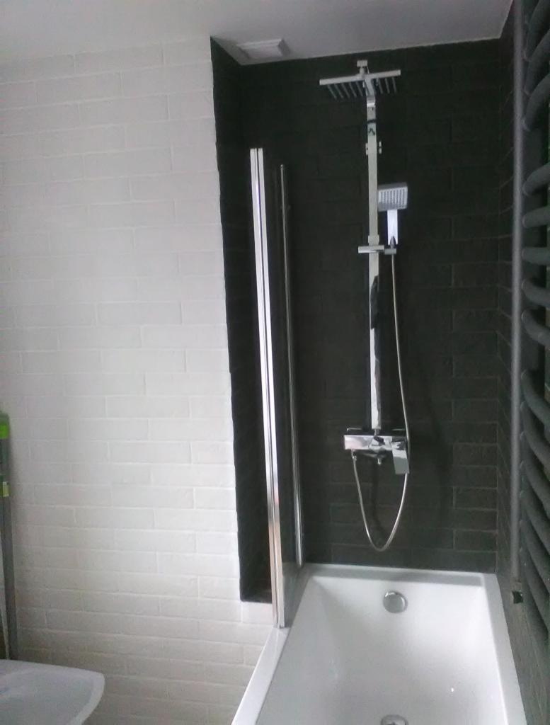 Nowoczesna łazienka z białą wanną, czarną ścianą z cegiełek i chromowaną deszczownicą z baterią, widoczna częściowo szklana osłona wanny.