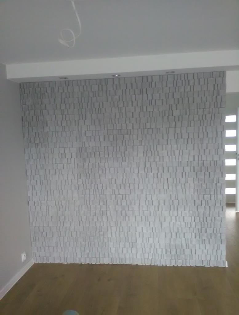 Ściana wykończona panelami 3D o nieregularnej fakturze, z widocznym kablem zwisającym z sufitu i drewnianą podłogą w nowocześnie urządzonym wnętrzu.