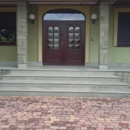 Wejście do domu z szarymi stopniami, kamienną okładziną ścian i brązowymi drzwiami wejściowymi z szybami.