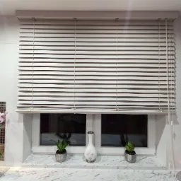 Jasne, poziome żaluzje w oknie, z dekoracjami na parapecie, w tle białe płytki i półki z ozdobami