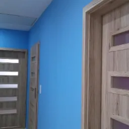 Dwie wewnętrzne drzwi z przeszkleniami, o fakturze jasnego drewna, na tle niebieskiej ściany w korytarzu z włazem na suficie.