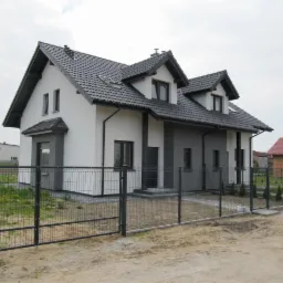 Nowoczesny dom jednorodzinny z dwuspadowym dachem krytym czarną dachówką, pomalowany na biało i szaro, otoczony metalowym ogrodzeniem panelowym na tle pochmurnego nieba.