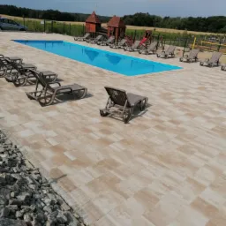 Beżowa kostka brukowa wokół basenu z leżakami, plac zabaw w tle. Nowoczesna aranżacja przestrzeni rekreacyjnej z dbałością o detale. Widok z góry na ułożoną kostkę.