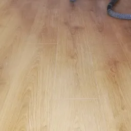 Jasnobrązowe panele podłogowe z widocznym wzorem drewna, w tle fragment węża odkurzacza przemysłowego.